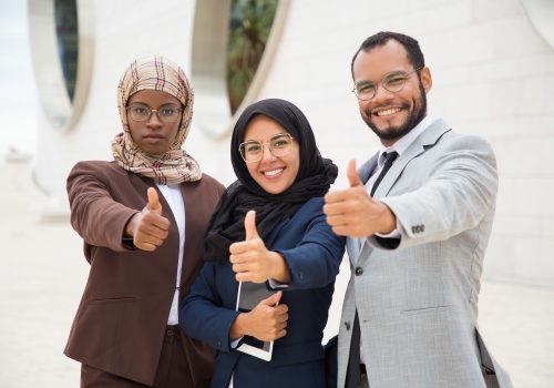 Multicultural business group posing and making like gesture. Businessman and Muslim businesswomen standing outdoors, showing thumb up and smiling at camera. Successful business team concept